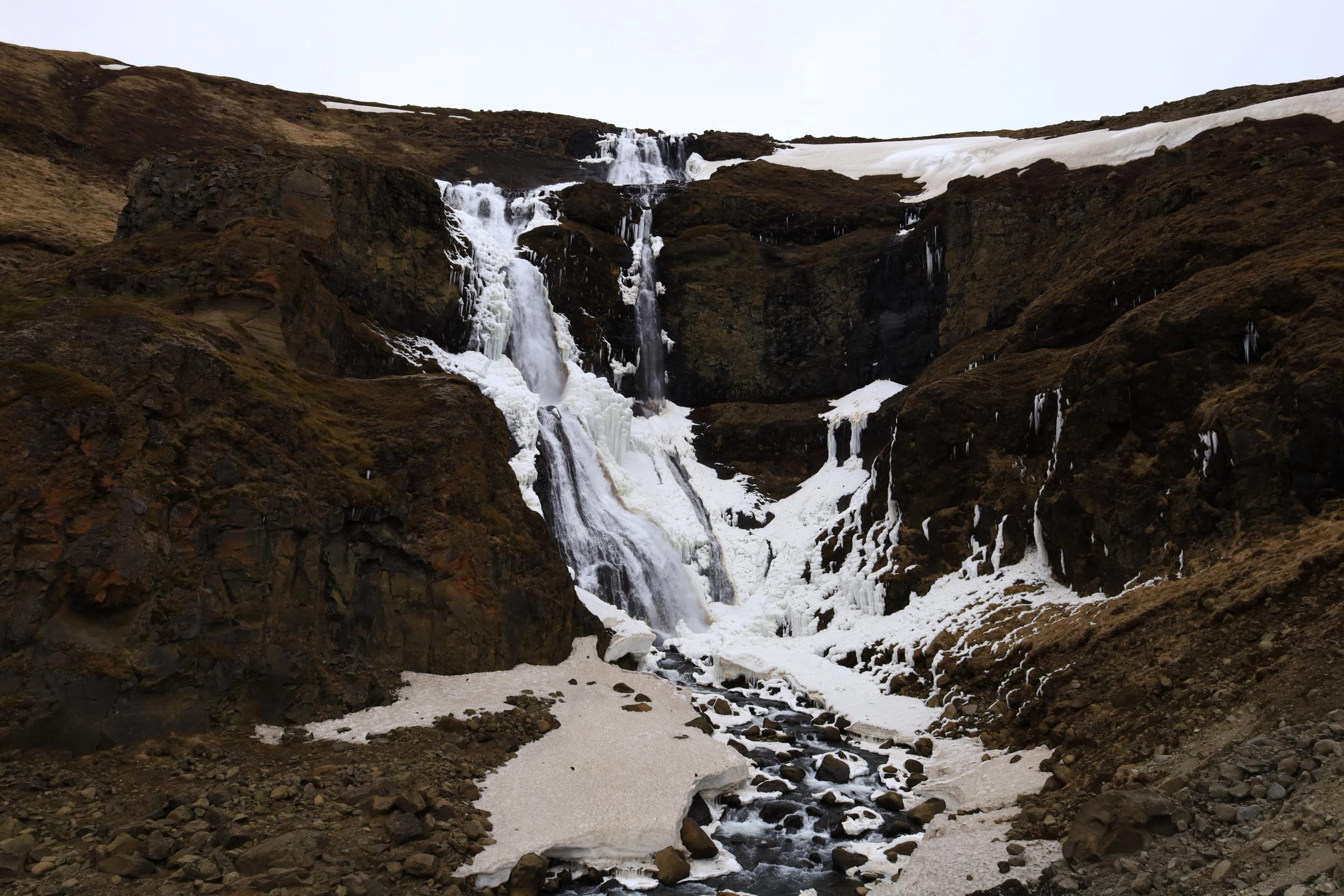 Partially frozen waterfall on a rocky, snow-covered hill.