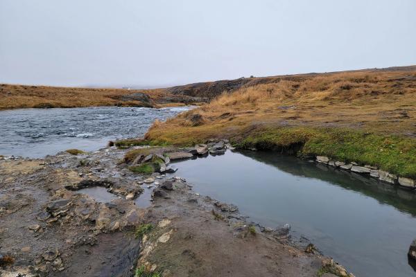 A fast-flowing river borders a calm pool on rocky, grassy terrain with golden hills under a grey sky.