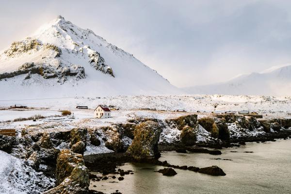 A white house on a snowy, rocky coast with a large snow-covered mountain in the background.
