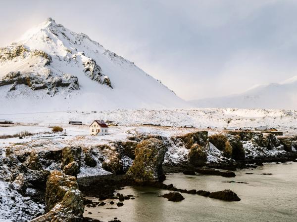 A small white house with a red roof is situated on a snowy, rocky coast, with an imposing snow-covered mountain behind it.
