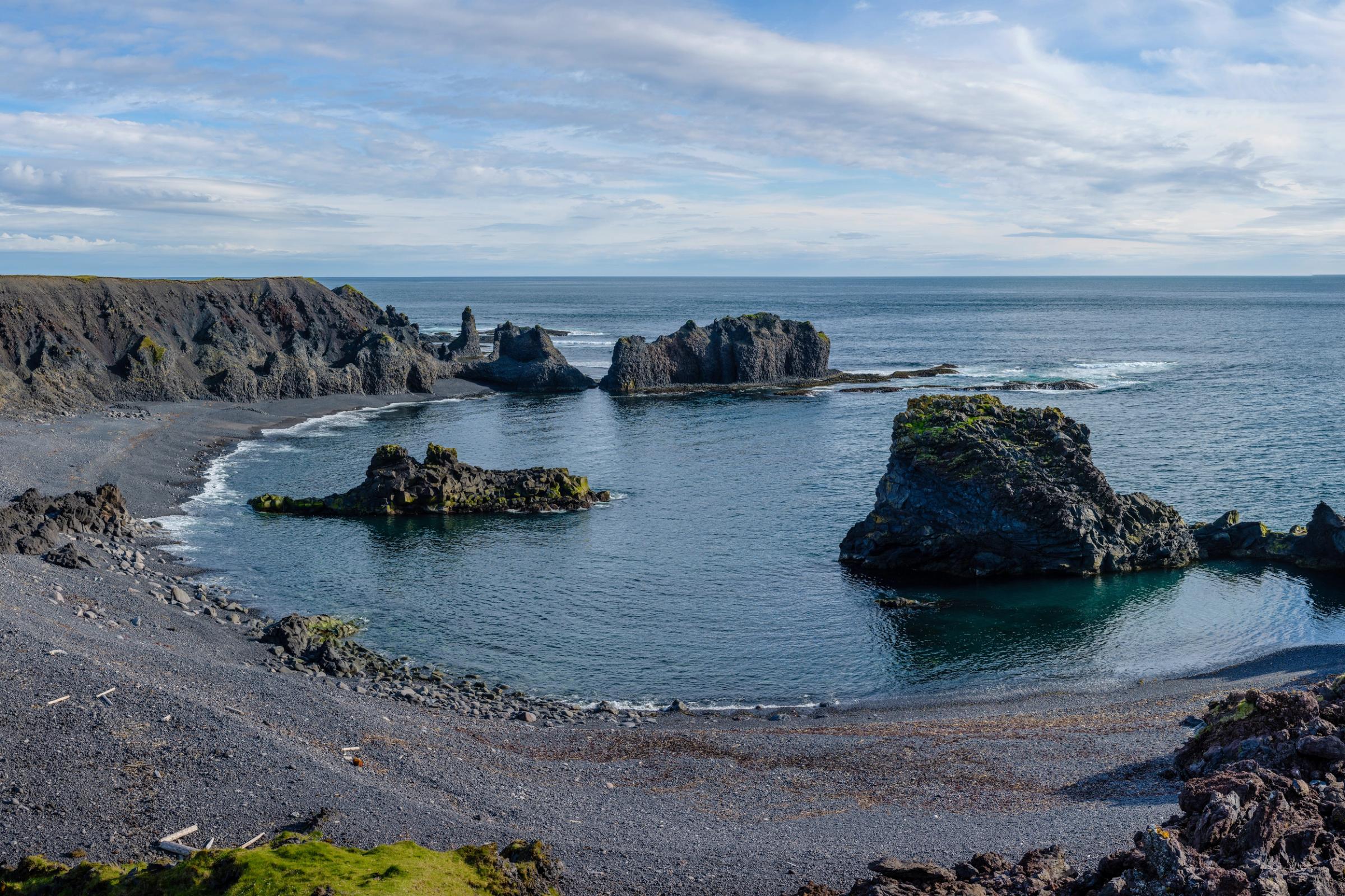 Panoramic view of a black sand beach with volcanic cliffs and sea stacks under a partly cloudy sky.