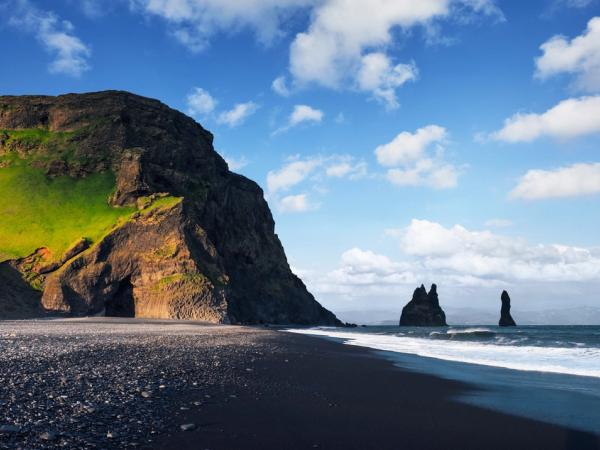 une plage de sable noir avec une montagne en arrière-plan et une grande formation rocheuse dans l'océan à Reynisfjara en Islande.