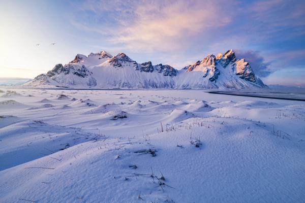 Snow-covered mountains with golden-lit peaks and snowy dunes under a colorful sunrise sky.