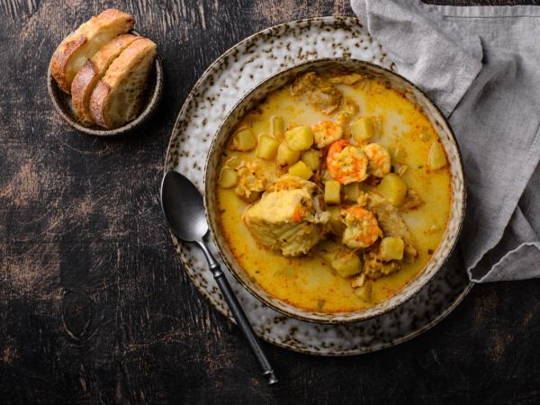 A bowl of yellow seafood stew with shrimp, potatoes, and fish, accompanied by sliced bread and a spoon on a dark surface.