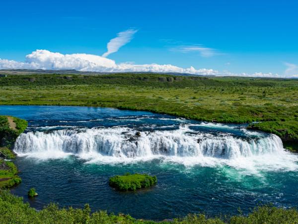 A wide waterfall flows into a blue river surrounded by green fields under a bright blue sky.