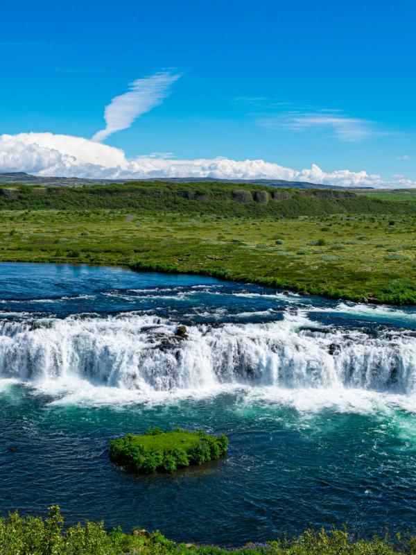 A wide waterfall flows through a lush green landscape under a bright blue sky.