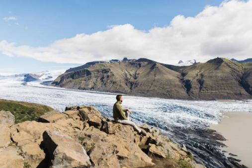 Un homme est assis au sommet d'une falaise rocheuse surplombant un glacier à Skaftafelljökull en Islande.