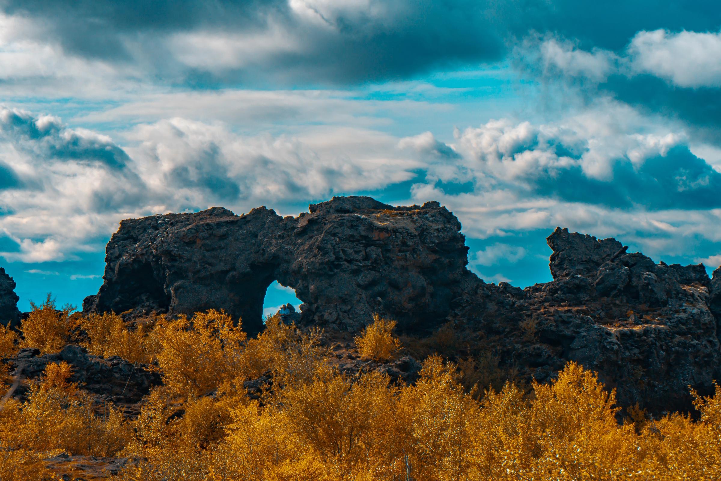 Oscuras rocas volcánicas con un arco natural, arbustos dorados y un cielo nublado dramático.