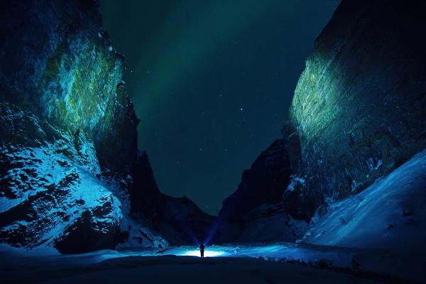 A person with a blue light illuminates a dark, snowy mountain valley under the Northern Lights.