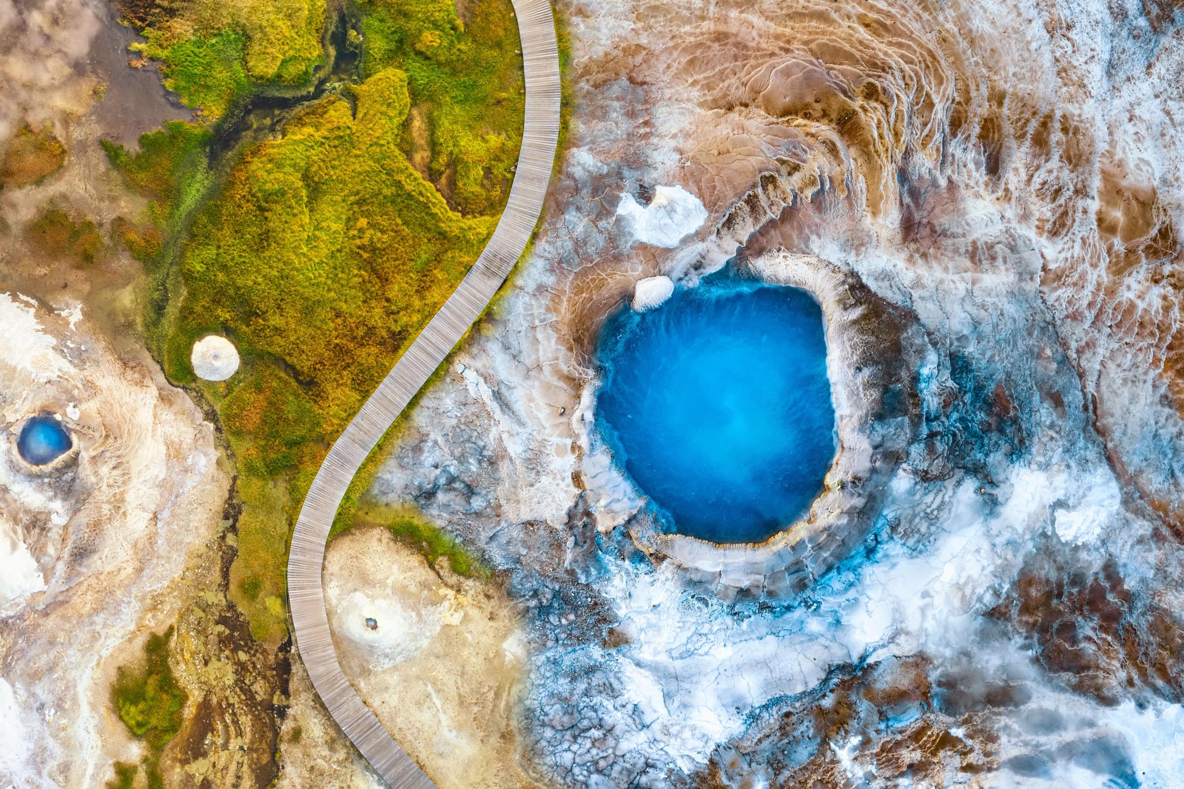 Vista aérea de una brillante piscina geotérmica de color azul, un sinuoso paseo de madera y coloridas formaciones minerales.