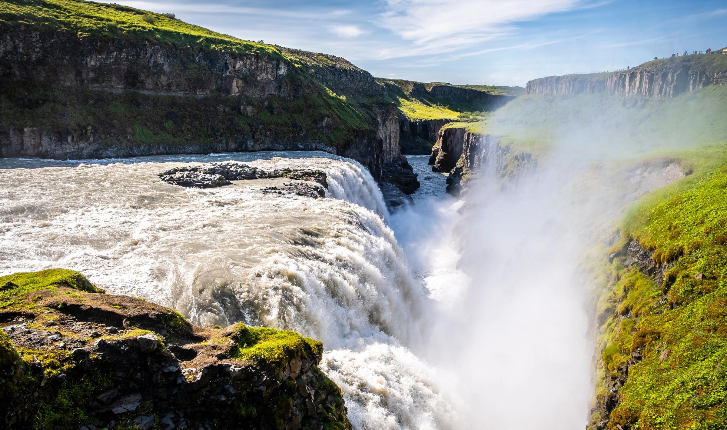 A wide waterfall plunges into a rocky canyon lined with green grass, creating a misty spray.