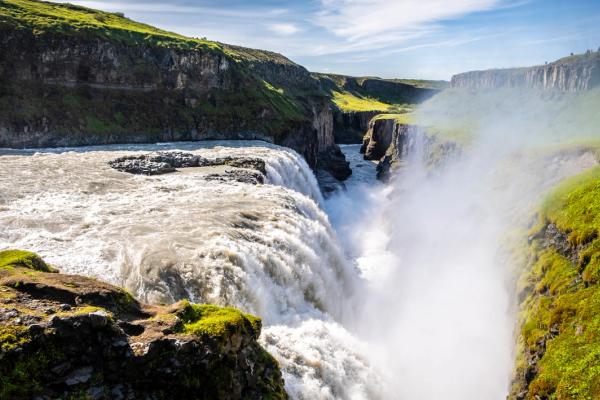 A wide waterfall plunges into a rocky canyon lined with green grass, creating a misty spray.