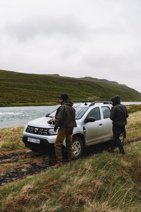 Dacia Duster parked on grass beside a river with two people standing nearby