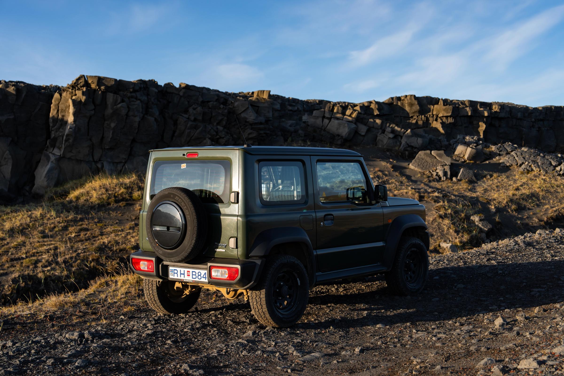 Suzuki Jimny mit Schaltgetriebe in der Dreiviertelansicht von hinten, mit Reserverad auf Schotter vor einer Felswand