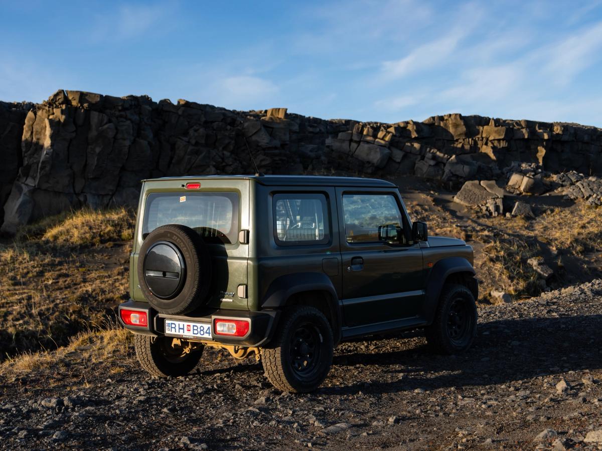Jimny Felswand hinten Suzuki Jimny mit Schaltgetriebe in der Dreiviertelansicht von hinten, mit Reserverad auf Schotter vor einer Felswand