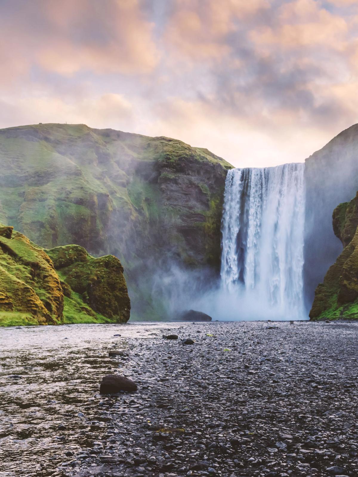 A powerful waterfall plunges between green hills into a rocky riverbed.