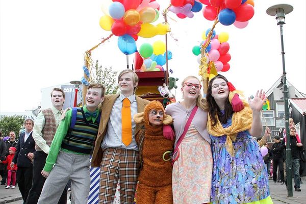 A joyful group of people in diverse costumes, including a monkey, smiling and waving under a canopy of colorful balloons at an outdoor event.