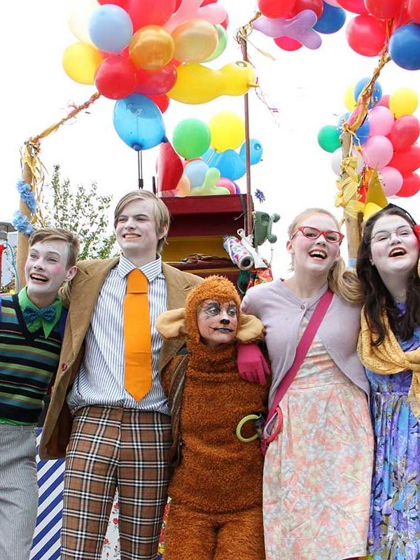 Iceland's national day A joyful group of people in diverse costumes, including a monkey, smiling and waving under a canopy of colorful balloons at an outdoor event.