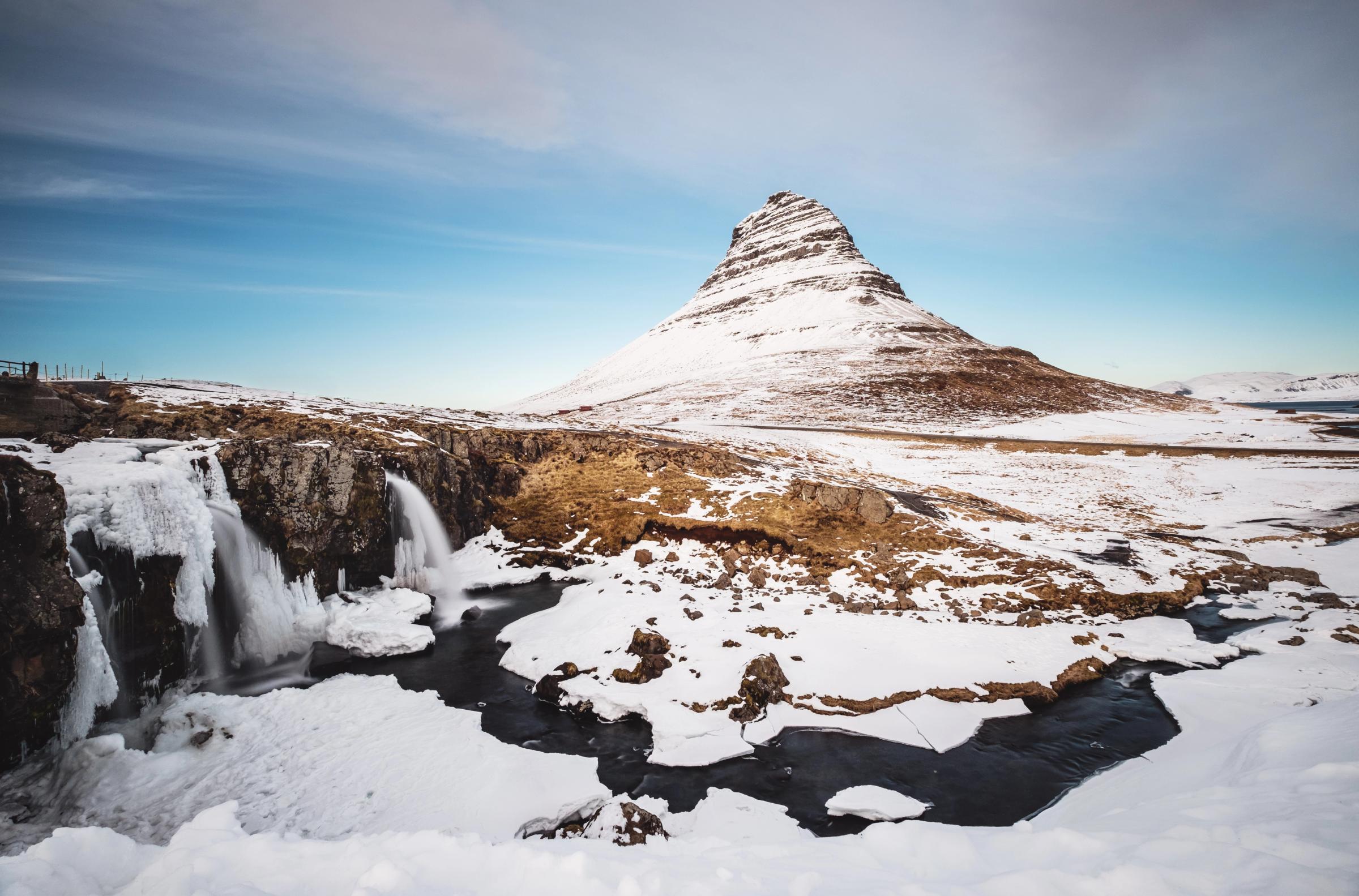 Snowy landscape featuring a conical mountain, a partially frozen waterfall, and a winding river under a blue sky.