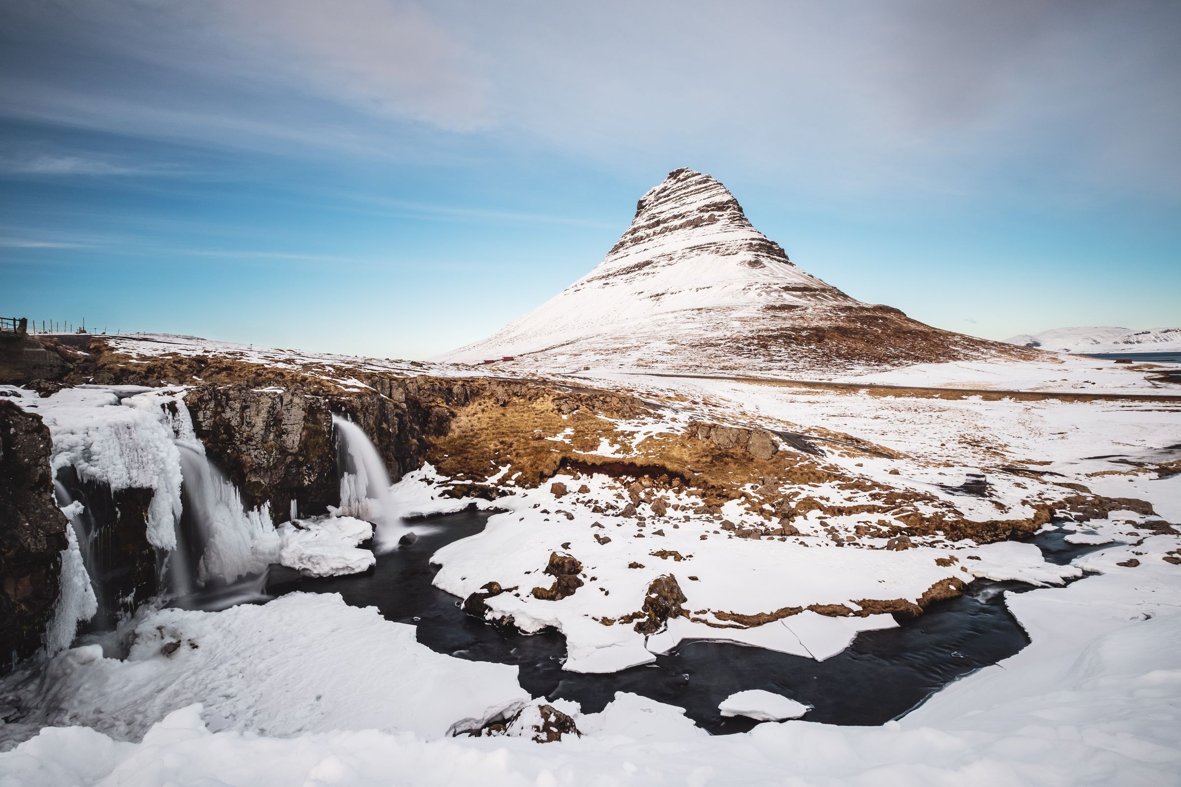 Snowy landscape featuring a conical mountain, a partially frozen waterfall, and a winding river under a blue sky.