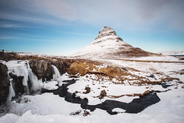 Snowy landscape featuring a conical mountain, a partially frozen waterfall, and a winding river under a blue sky.