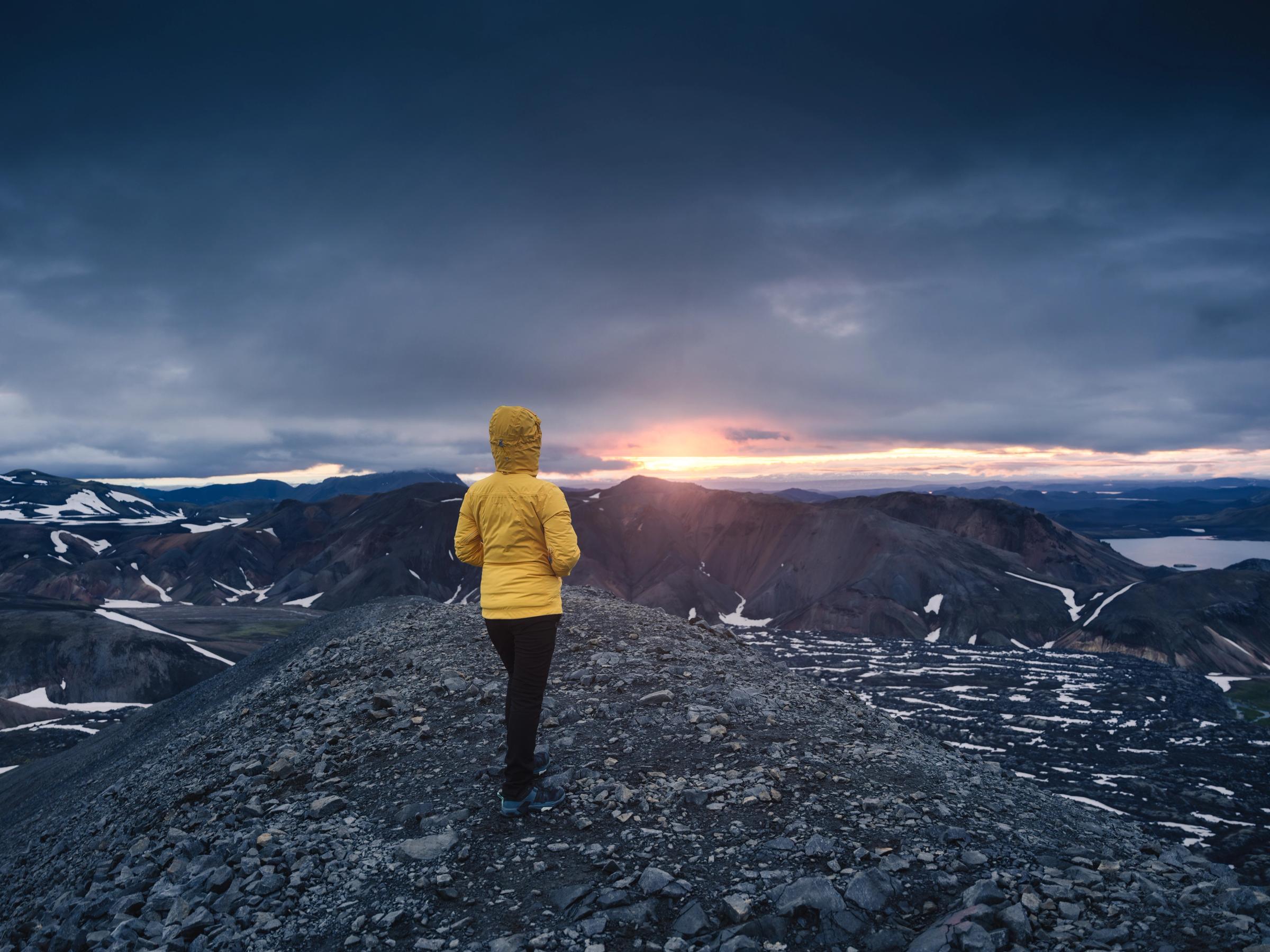 Una persona con una chaqueta amarilla está de pie en una cresta rocosa de la montaña, contemplando un paisaje vasto y escarpado con un lago bajo un cielo dramático y nublado al atardecer.