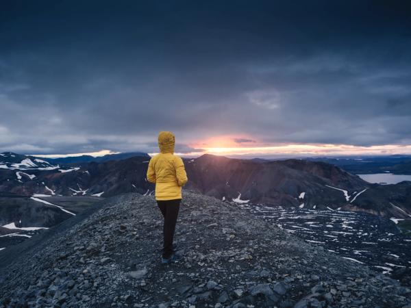 Una persona con una chaqueta amarilla está de pie en una cresta rocosa de la montaña, contemplando un paisaje vasto y escarpado con un lago bajo un cielo dramático y nublado al atardecer.