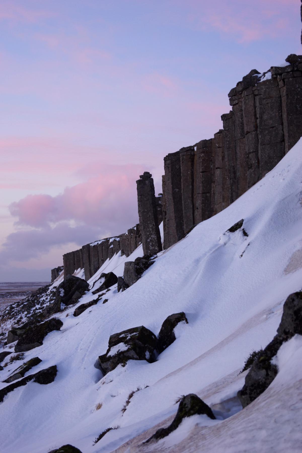 Basalt column cliffs rise from a snowy slope under a pink and purple twilight sky.