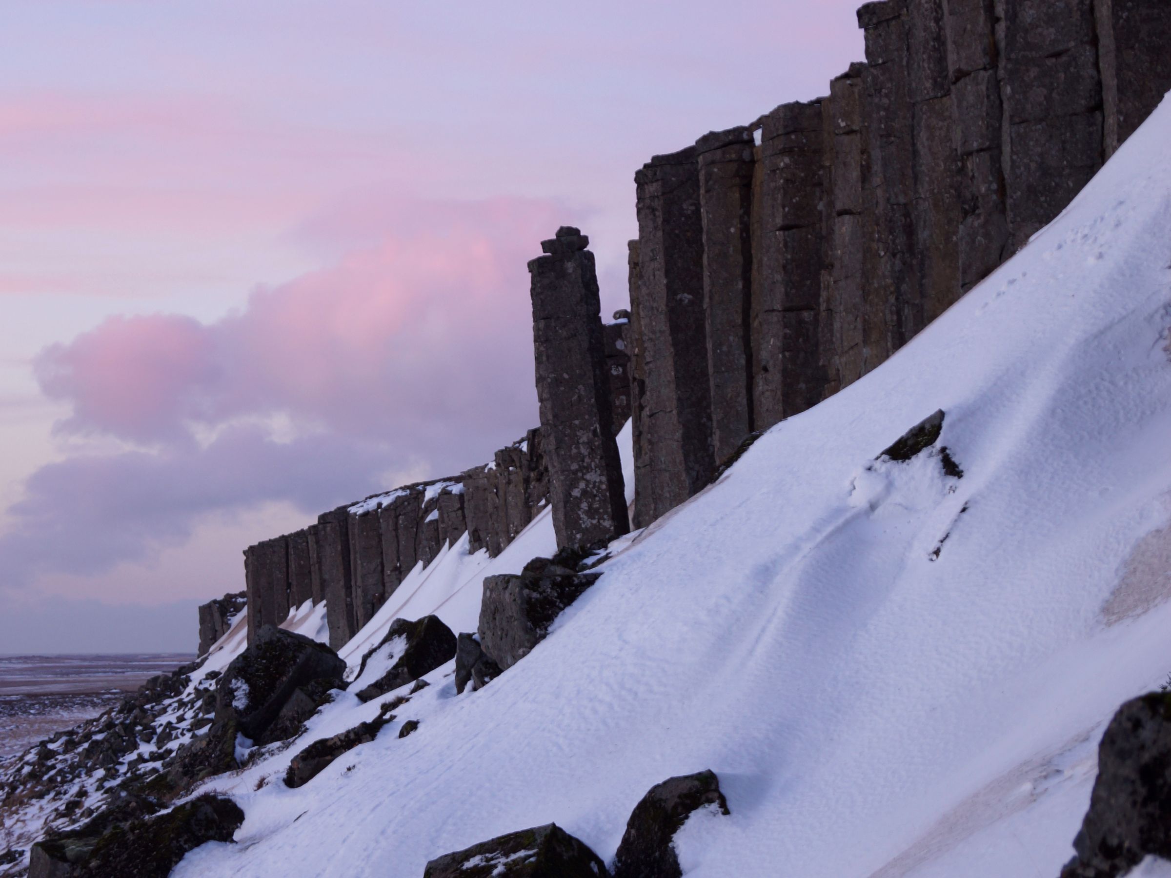 Basalt column cliffs rise from a snowy slope under a pink and purple twilight sky.