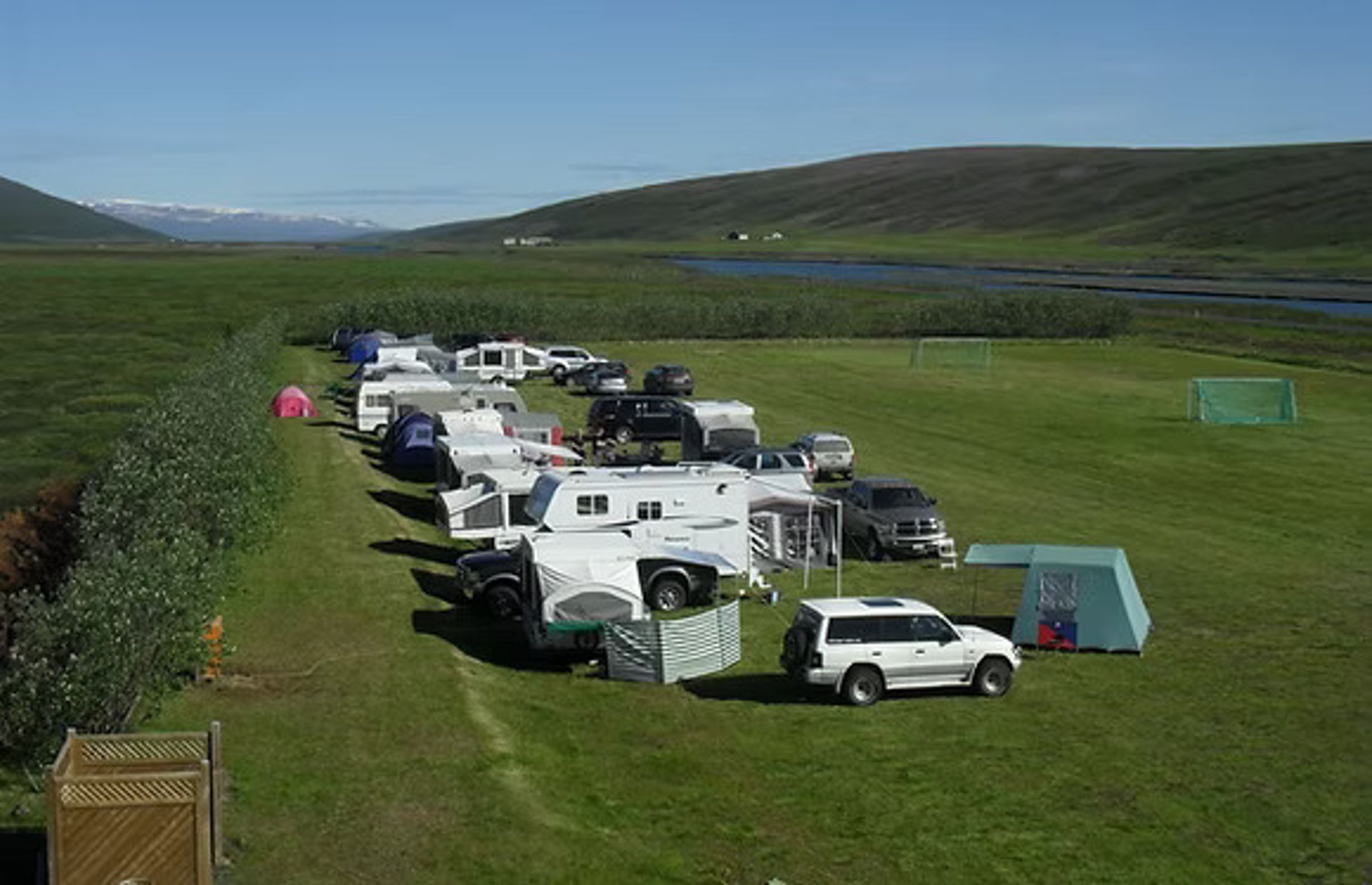 A campground with RVs, trailers, and tents on a green field, backed by a river, hills, and distant mountains.