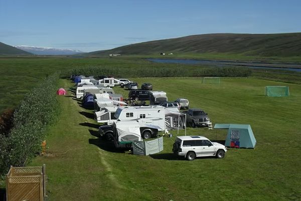 A campground with RVs, trailers, and tents on a green field, backed by a river, hills, and distant mountains.
