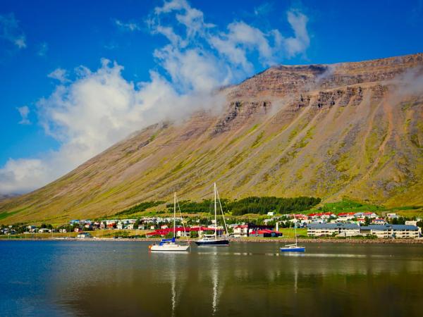 Coastal town with sailboats in a fjord, backed by a cloud-covered mountain under a blue sky.