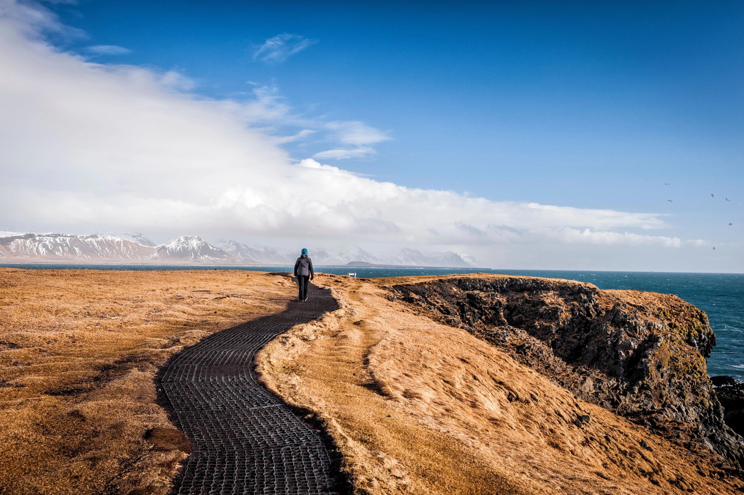 A person walks on a winding path along a coastal cliff overlooking the sea, with distant snow-capped mountains.