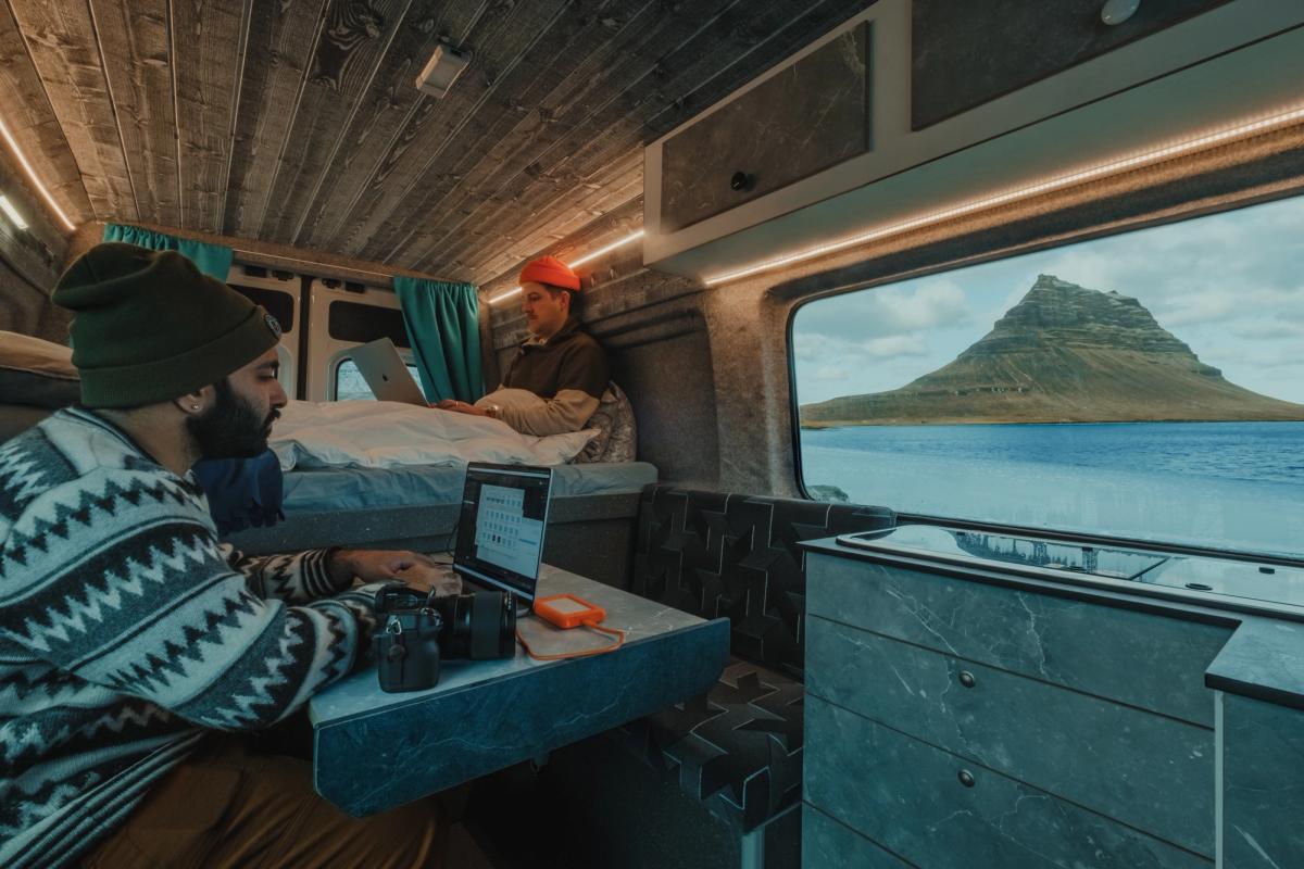 Dos personas con portátiles en la mesa y una cama al fondo junto a una gran ventana lateral