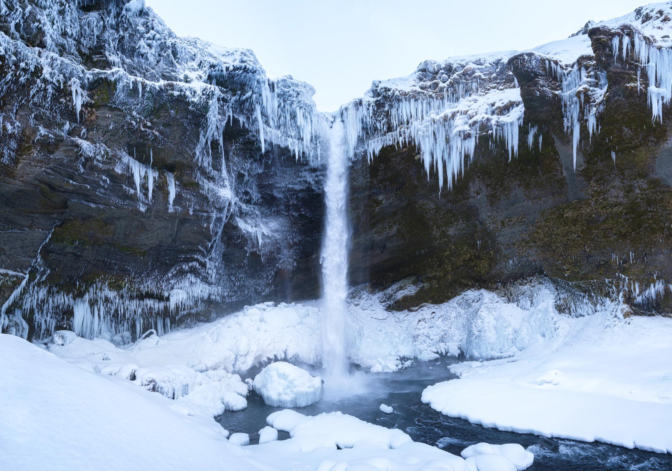 A tall waterfall cascades into a snowy, icy riverbed, flanked by snow-covered cliffs with long icicles.