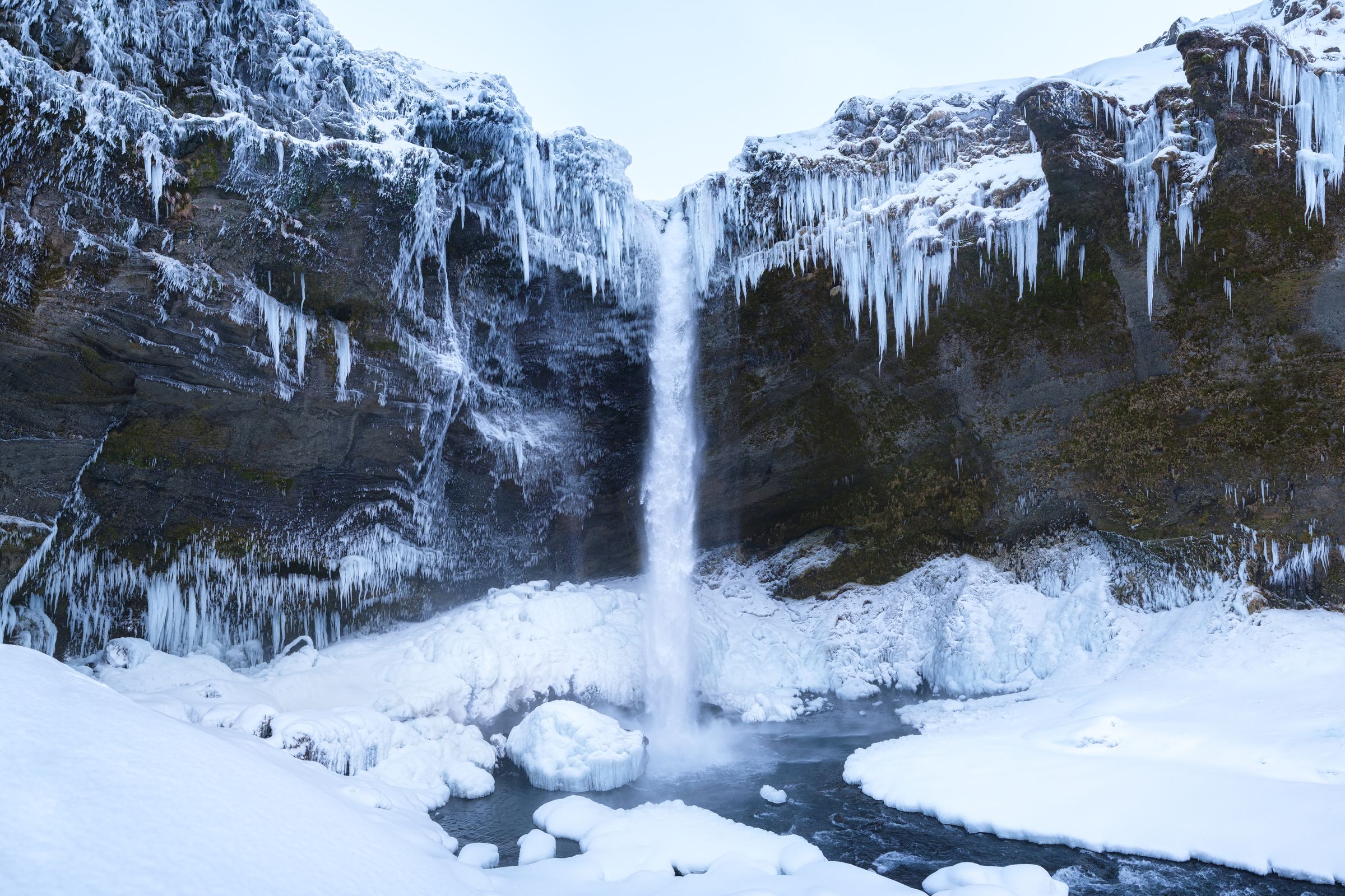 A tall waterfall cascades into a snowy, icy riverbed, flanked by snow-covered cliffs with long icicles.