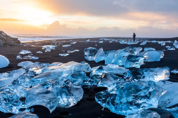 Black sand beach scattered with clear ice chunks, a person in the distance, and a warm sunset sky.