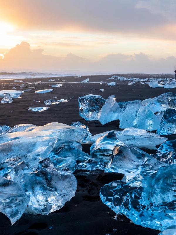 Glistening ice chunks on a black sand beach at sunset, with a person in the distance.