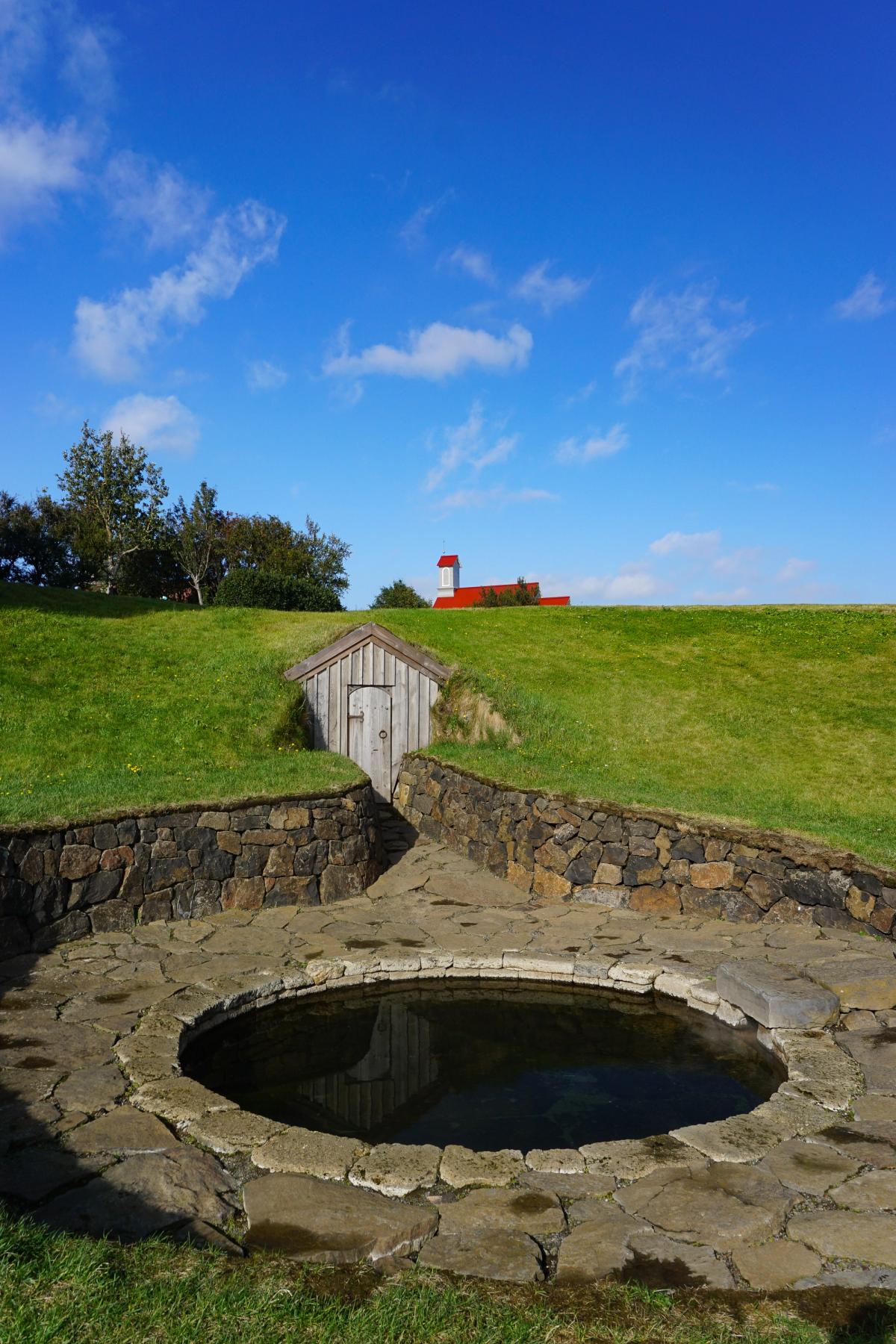 a stone wall surrounds a pool of water with a red building in the background at Snorralaug in Reykholt iceland.