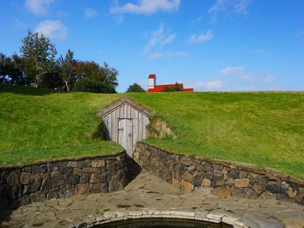 a stone wall surrounds a pool of water with a red building in the background at Snorralaug in Reykholt iceland.