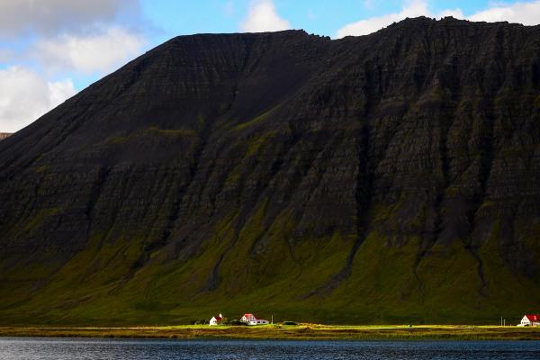 A dark, rocky mountain with green lower slopes overlooks a body of water, with small red-roofed houses along its shore.