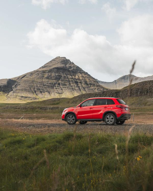 Red SUV parked on a dirt road in a grassy landscape with a large, layered mountain under a cloudy sky.