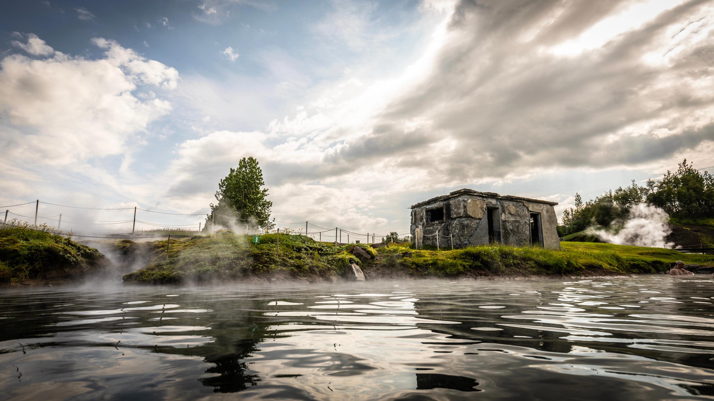 A steamy geothermal landscape with a dilapidated stone hut and a lone tree on the bank.