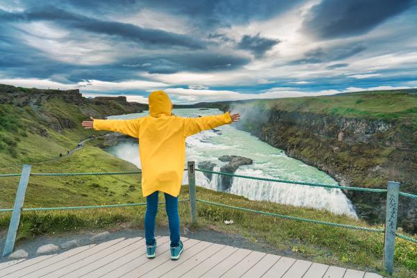 A person in a yellow raincoat with arms outstretched overlooks a powerful waterfall in a green canyon under a cloudy sky.