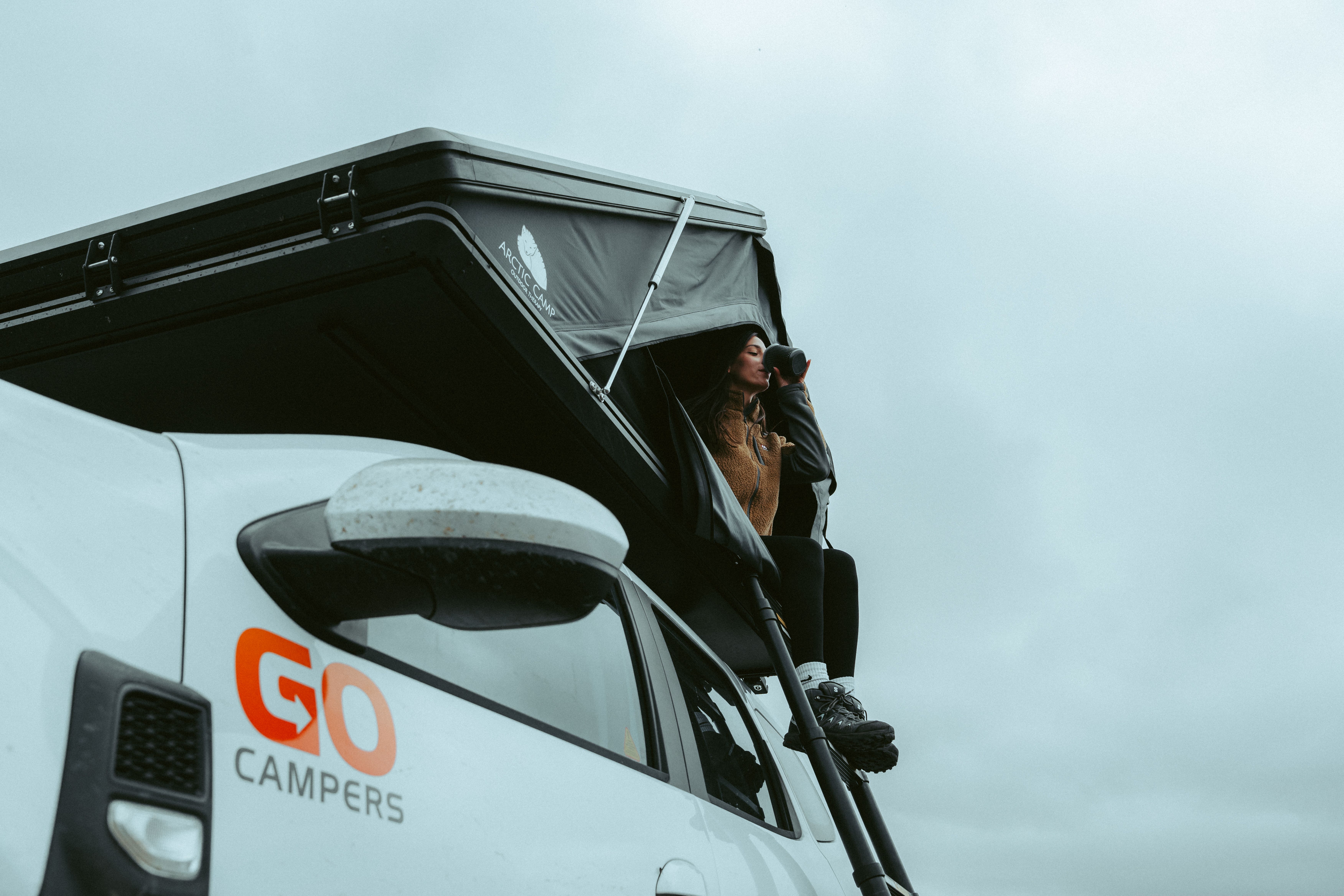 Person sitting at the rooftop tent entrance with a mug, above the Go Campers door logo