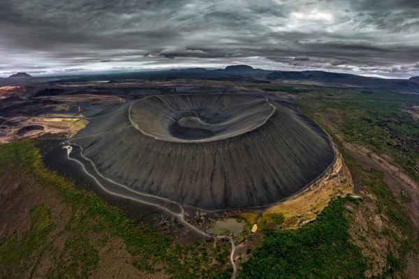 Aerial view of a dark, conical volcanic crater with terraced inner walls and a path leading to its rim, surrounded by dark earth and green patches under a cloudy sky.
