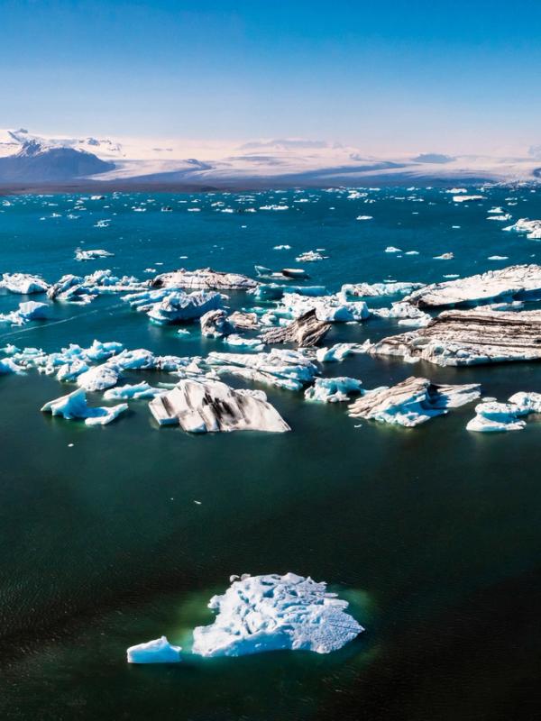 Panoramic view of a glacial lagoon filled with icebergs, dark shores, and snow-capped mountains.