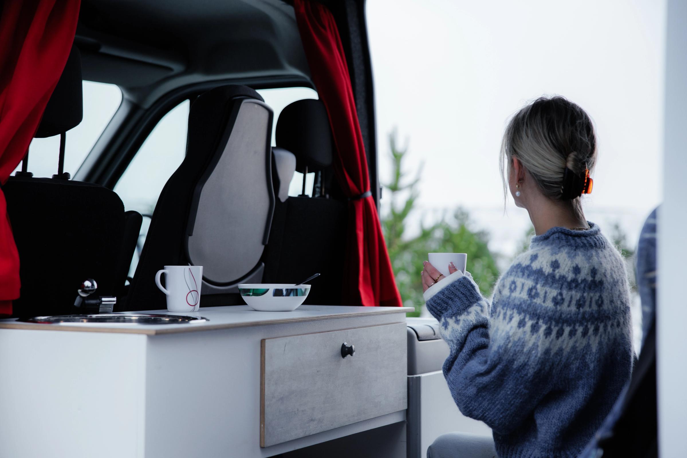 Frau mit einer Tasse in der Hand neben der Küchenzeile, die aus einem großen Camper für fünf Personen nach draußen schaut
