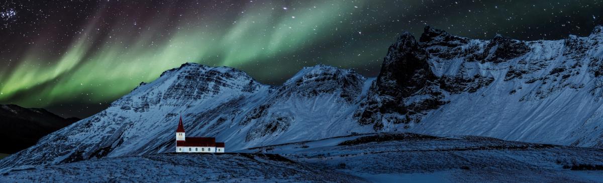 A white church with a red roof stands in a snowy mountain landscape under a starry sky with green aurora borealis.