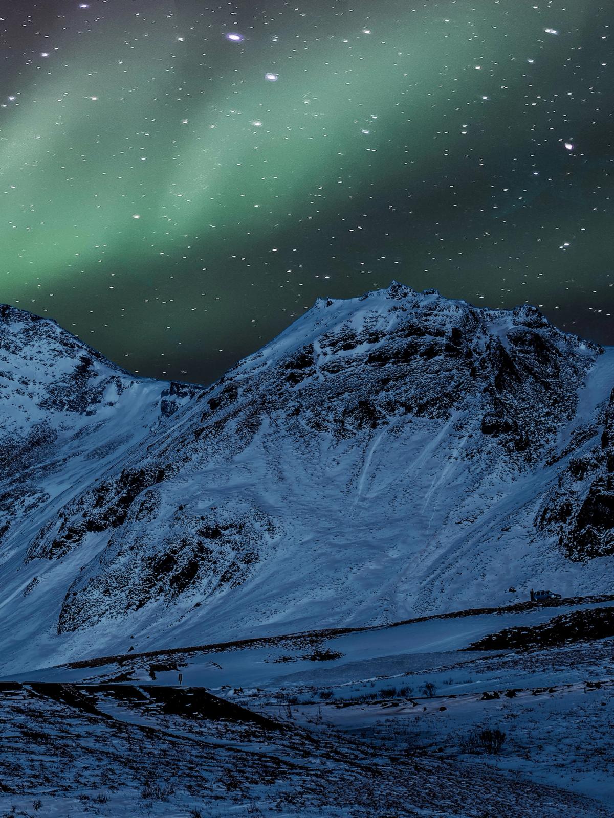 A white church with a red roof stands in a snowy mountain landscape under a starry sky with green aurora borealis.